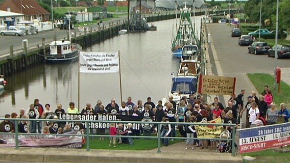 Eine Gruppe Menschen steht protestierend vor dem Hafenbecken von Friedrichskoog. © NDR Foto: Screenshot