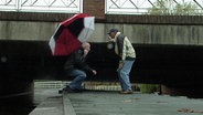 Zwei Männer bücken sich auf einem Steg stehend, um unter einer darüber verlaufenden Brücke hindurchzukommen. © NDR Foto: Screenshot