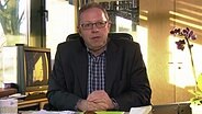 Niels Schmidt, der Bürgermeister von Wedel, sitzt hinter seinem Schreibtisch, auf dem eine Orchidee und ein Wackel-Dackel stehen. © NDR Foto: Screenshot