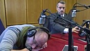 Ein Radiomoderator liegt eingeschlafen auf dem Tisch, Johannes Schlüter blickt ihn fragend an. © NDR Foto: Screenshot
