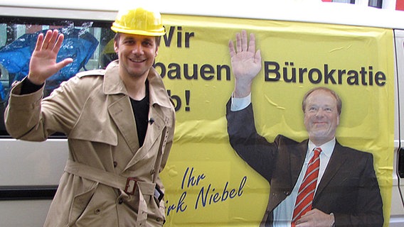 Tobias Schlegl winkt neben einem Plakat von Dirk Niebel in die Kamera.  