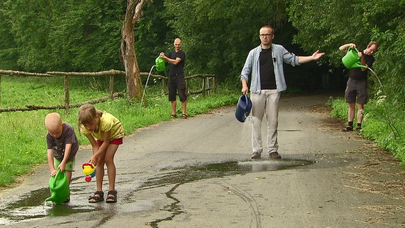 Drei Männer stehen auf einem geteerten Radweg und gießen Wasser aus Kannen aus. © NDR Foto: Screenshot