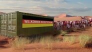 In der Wüste steht ein Container mit deutscher Flagge, davor eine lange Schlange Menschen.  