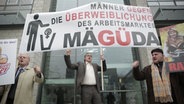 Johannes Schlüter steht protestierend unter einem Plakat mit der Aufschrift MÄGÜDA: Männer gegen die Überweiblichung des Arbeitsmarktes.  