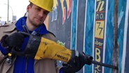 Tobias Schlegl, als Bauarbeiter verkleidet, hält einen Presslufthammer an die Berliner East Side Gallery. © NDR Foto: Julian Amershi