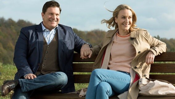 Ein Mann (links) und eine Frau (rechts) sitzen lächelnd nebeneinander auf einer Bank im Grünen. Sie halten ihre Hände. © © ARD/Volker Roloff, honorarfrei 