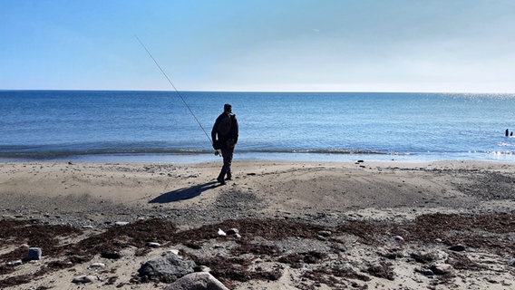 Ein Angler von hinten mit einer Angel in der linken Hand steht am Strand und guckt auf das Meer.  Foto: Martin Möller