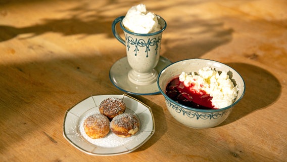 Auf einem Tisch stehen ein Teller mit Pförtchen, eine Schale Milchreis mit Kirschen und ein Becher Kakao mit Sahne. © NDR/Doclights GmbH 