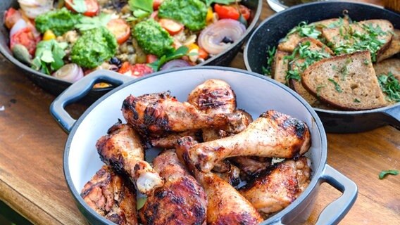 Marinierte und gegrillte Hähnchenkeulen in einer Schüssel. In weiteren Gefäßen sind mediterranes Grillgemüse sowie geröstete Brotscheiben © NDR 