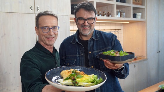 Tarik Rose und Dr. Matthias Riedl stehen in einer Küche und halten zwei Teller mit Ofengerichten in die Kamera. © NDR Foto: Florian Kruck