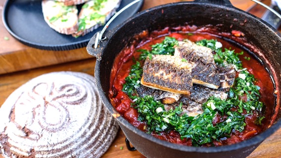 Meeräsche auf orientalischem Tomaten-Ragout mit Petersilien-Pesto in einem Topf. Daneben liegen ein Laib Bauernbrot sowie gewürzte Butter-Stullen. © NDR 