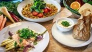 Auf einem Tisch stehen ein Teller mit Linsensalat, Spargel und Brokkoli mit Lachsforelle sowie Quarkcreme und Rhabarber in einem Pergament-Päckchen. © NDR Foto: Tarik Rose