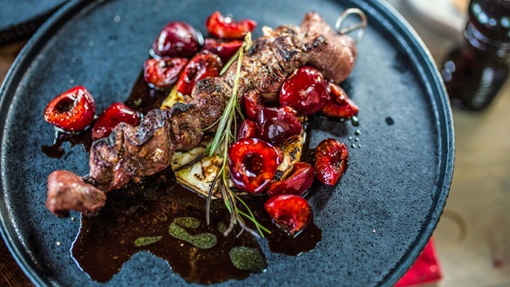 Ein Rehfilet-Spieß auf gegrillter Sellerie mit Kirsch-Chutney angerichtet auf einem blauen Essteller © NDR Foto: Tarik Rose