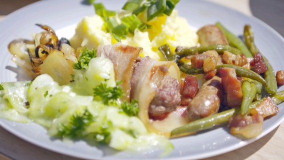 Rindfleisch im Speckmantel und Merguez-Würste mit Kartoffelpüree und Speckbohnen an Petersilien-Gurkensalat auf einem Teller angerichtet © NDR Foto: Tarik Rose