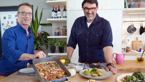 Fernsehkoch Tarik Rose und Ernährungs-Doc Matthias Riedl stehen in einer Küche und zeigen einen Teller mit Fischfrikadellen und eine Auflaufform mit Apple Crumble. © NDR/dmfilm/Florian Kruck 