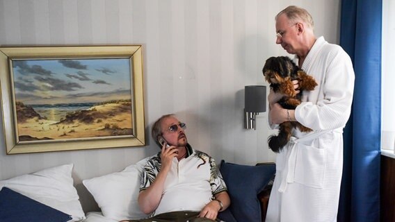 Szenenbild aus der Comedy-Serie "Jennifer - Sehnsucht nach was Besseres": Dietmar (Olli Dittrich, li.) sitzt auf dem Bett und blickt zu Hans (Ludger Pistor, re.) hoch, der einen Hund auf dem Arm hat. © NDR/Marion von der Mehden 