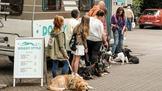 Szenenbild aus der Comedy-Serie "Jennifer - Sehnsucht nach was Besseres": Hundebesitzer stehen vor Jennifers Hundesalon Doggy Style Schlange. © NDR/Marion von der Mehden 