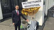 Michel Abdollahi steht neben einem Klappstuhl, über dem ein Schild hängt: Weltrettungsbeichtstuhl. © NDR / Stefan Mühlenhoff 