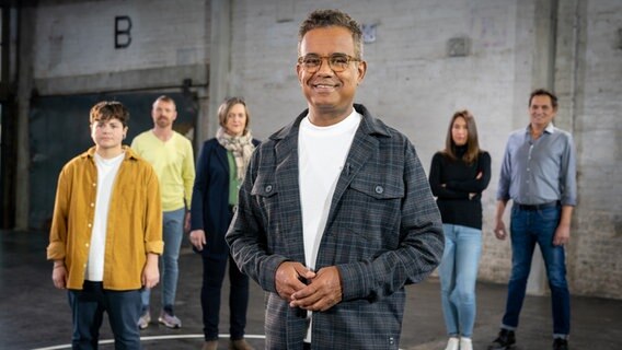 Ein Mann steht im Vordergrund, eine Gruppe von Menschen steht im Hintergrund in einer Reihe © NDR 