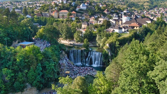Viele Menschen stehen vor zwei großen Wasserfällen, dahinter liegt eine Ortschaft. © NDR/micafilm/Dado Ruvić, honorarfrei Foto: Dado Ruvić