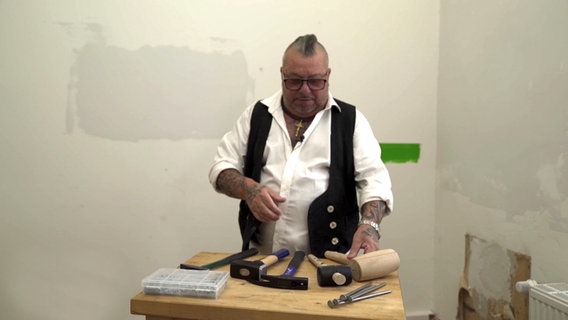 Ein Mann steht vor einem Tisch, auf dem verschiedene Hämmer liegen. © NDR/nonfictionplanet 