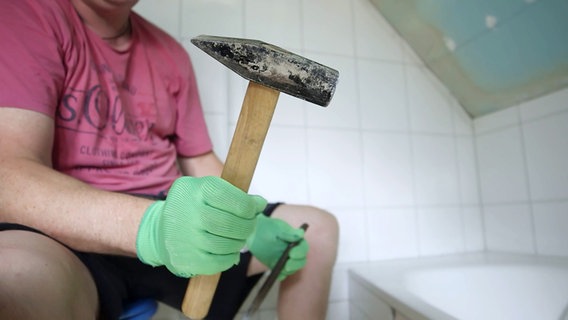 Ein Mann sitzt in seinem Badezimmer und zeigt sein Werkzeug für den Abriss: Hammer und Meißel. © NDR/nonfictionplanet 