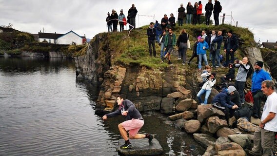 Ein junger Mann flippt Steine über das Wasser. Hinter ihm stehen einige Zuschauer. © NDR/nonfictionplanet/Michael Mc Glinn 