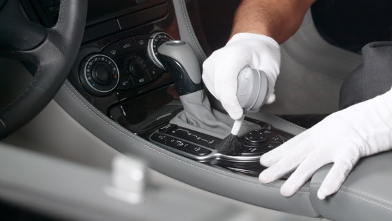 Mann mit mit Stoffhandschuhen reinigt die Mittelkonsole eines Autos mit einem Pinsel. © fotolia Foto: Günter Menzl