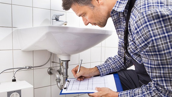 Handwerker kniet vor einem Waschbecken und macht sich Notizen. © Fotolia.com Foto: apops