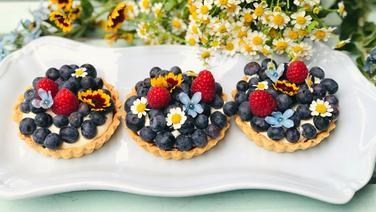 Frucht-Tartelettes mit Blaubeeren und essbaren Blüten auf einer Platte serviert. © NDR 