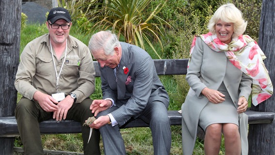 Prince Charles und Camilla sitzen auf eine Bank und lachen, weil Charles eine agile kleine Echse in der Hand hält. © picture-alliance/Schwörer Pressefoto Foto: Kai Schwörer