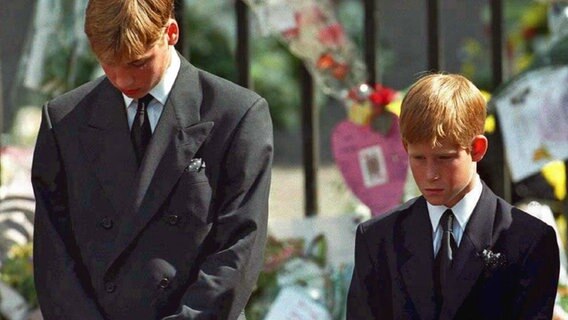 Prinz Harry steht mit gefallteten Händen und gesenktem Haupt neben seinem Bruder Prinz William. © picture alliance / dpa 