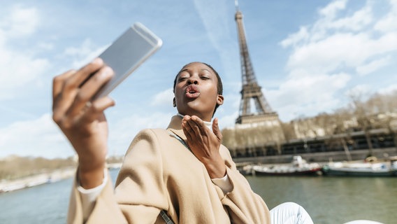 Wir sehen eine junge Frau in Paris, die mit ihrem Smartphone ein Selfie macht. Hinter ihr steht der Eiffelturm. © NDR 