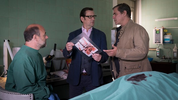 Drei Männer in einem Raum in der Pathologie schauen sich an. Der Mann in der Mitte hält einen blutigen, zerrissenen Zettel in der Hand. © NDR/ARD/Sandra Hoever Foto: Sandra Hoever