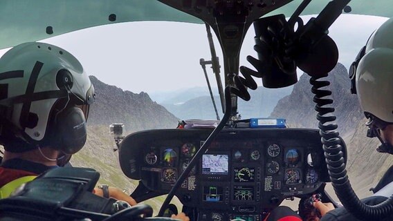 Blick vom hinteren Teil eines Helikopters nach draußen auf die Berge. Vorn sitzen zwei Männer von der Bergrettung. © NDR/BR 
