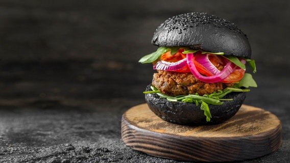 Ein Burger mit schwarzen Brötchenhälften, Tomaten, Ruccola und roten Zwiebeln liegt auf einem Brett. © picture alliance / Zoonar | Oleksandr Latkun Foto: Oleksandr Latkun