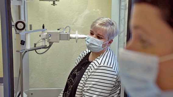 Patienten mit Post-Covid-Symptomen sitzt in einer Kabine, in der die Lungenfunktion gemessen wird. Am Anschnitt ist eine Frau mit OP-Maske zu sehen.  
