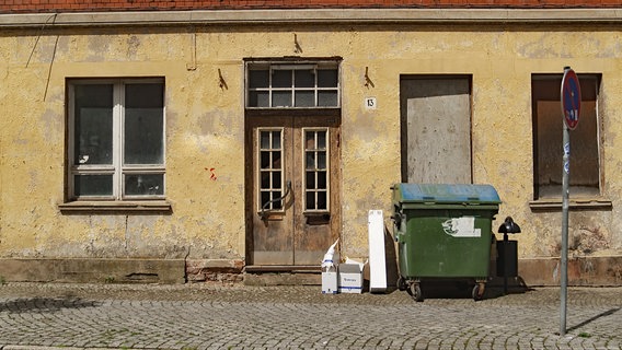 Heruntergekommene Fassade eines leerstehenden Hauses. Zwei von drei Fenstern sind mit Brettern verschlossen. Davor stehen ein großer Müllcontainer sowie Kartons. © MDR/Hoferichter & Jacobs 