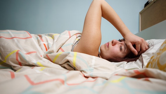 Eine Frau liegt im Bett und greift sich an den Kopf. Sie sieht erschöpft aus. © Colourbox Foto: -