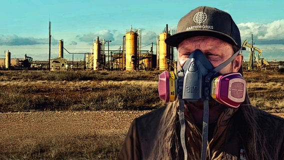 Ein Mann mit Atemmaske aus Gummi mit Filterkammern links und rechts vor einer flachen Landschaft mit Gas-Silos in den USA. © NDR | HTTV Produktion / Michael Höft 