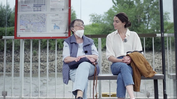 Eine ältere Frau und und eine junge sitzen auf der Bank eines Fähranleger-Wartehäuschens. © NDR 