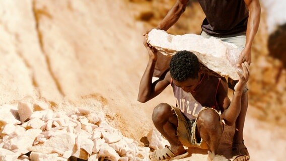 Einem jungen Mann wird in einem Abbaugebiet für Rosenquarz ein schwerer Stein auf die Schultern geladen. © NDR 