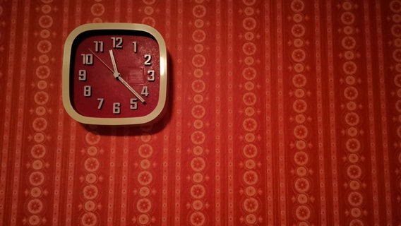 Eine Uhr mit rotem Hintergrund hängt an einer Wand mit roter Tapete. © NDR/Jasmin Kabus-Bardoux 