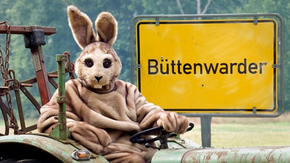 Als Hase Verkleideter auf Traktor sitzend, dahinter Ortsschild von Büttenwarder (Montage) © NDR/Nicolas Maack 