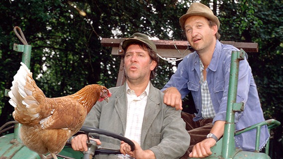 Die Bauern Brakelmann (Jan Fedder) und Tönnsen (Peter-Heinrich Brix) sitzen auf Trecker mit Huhn auf der Motorhaube. © NDR/I. Walther Foto: I. Walther