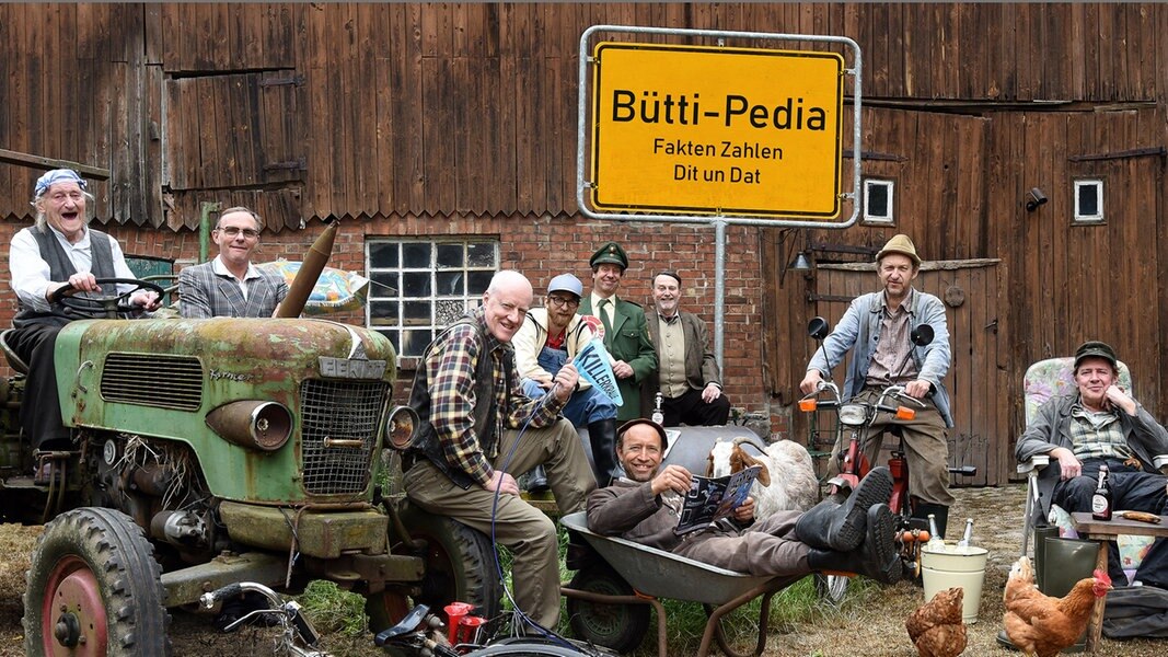 Bütti-Pedia: Menschen aus Büttenwarder stehen, sitzen um ein Ortsschild mit der Aufschrift: Bütti-Pedia - Fakten - Zahlen - Dit un Dat.