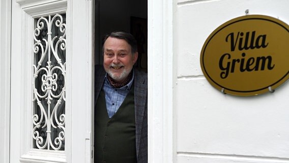 Ein Mann (Günther Griem, Bürgermeister von Büttenwarder) guckt aus der Haustür einer weißen Villa. © NDR/Nico Maack 