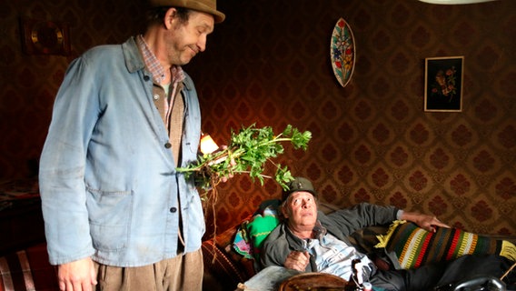 Szenenbild der 62. Büttenwarder-Folge "Haggnschuss": Adsche bringt dem verletzten Brakelmann Blumen mit. © NDR/Nicolas Maack Foto: Nicolas Maack