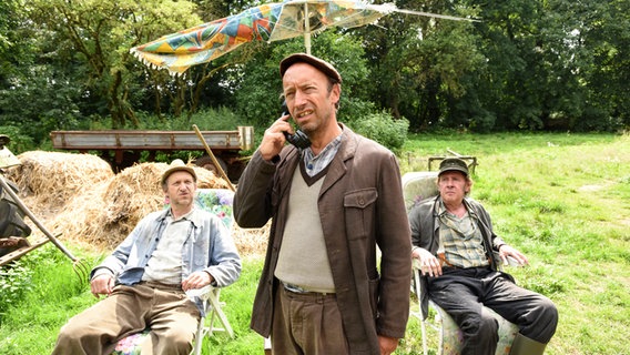 Szenenbild aus der 72. Büttenwarder-Folge "Oh!": Adsche und Brakelmann sitzen im Garten während Kuno telefoniert. Szenenbild aus der 72. Büttenwarder-Folge "Oh!": Adsche und Brakelmann sitzen im Garten während Kuno telefoniert. © Polyphon / Uwe Ernst für NDR Foto: Uwe Ernst