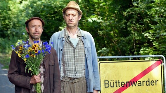 Kuno hält einen Blumenstrauß in der Hand und steht mit Adsche an Ortsschild. Kuno hält einen Blumenstrauß in der Hand und steht mit Adsche an Ortsschild. © NDR/Nico Maack Foto: Nico Maack